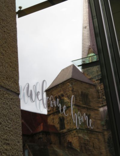 welcome-home-Schriftzug auf Fenster, in dem sich ein Teil des Doms spiegelt